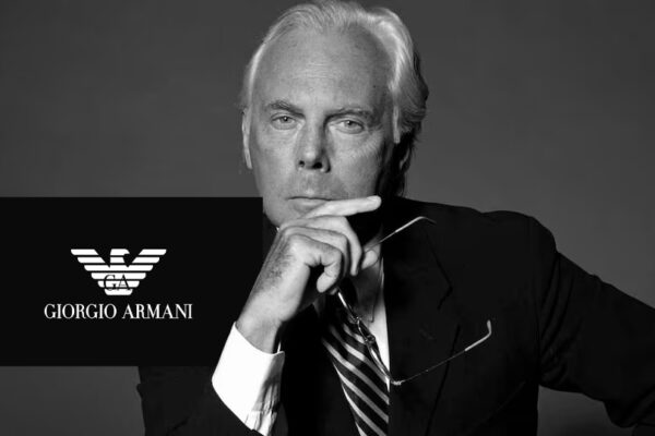 Black-and-white portrait of Giorgio Armani in his Milan studio, wearing a simple dark crew-neck and smiling, reflecting the minimalist elegance that defined his brand.