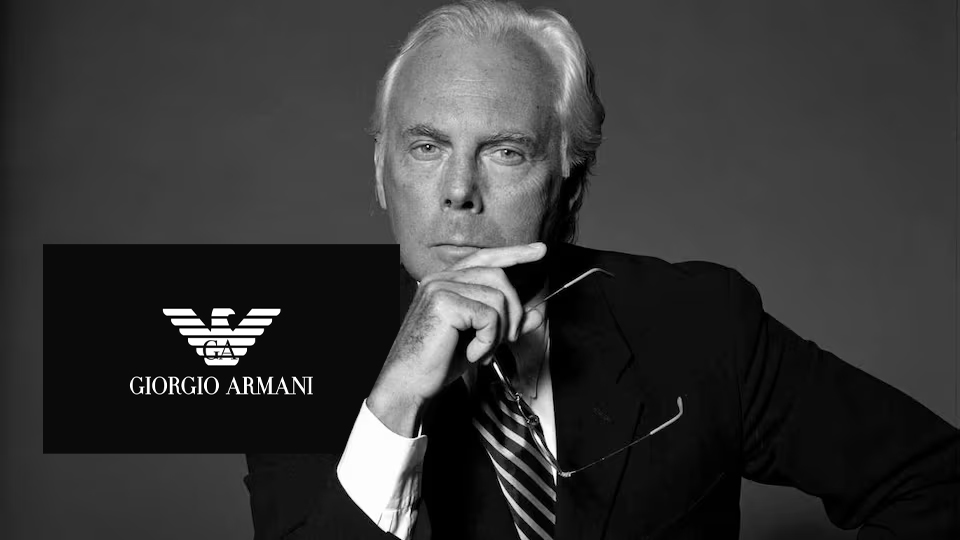 Black-and-white portrait of Giorgio Armani in his Milan studio, wearing a simple dark crew-neck and smiling, reflecting the minimalist elegance that defined his brand.